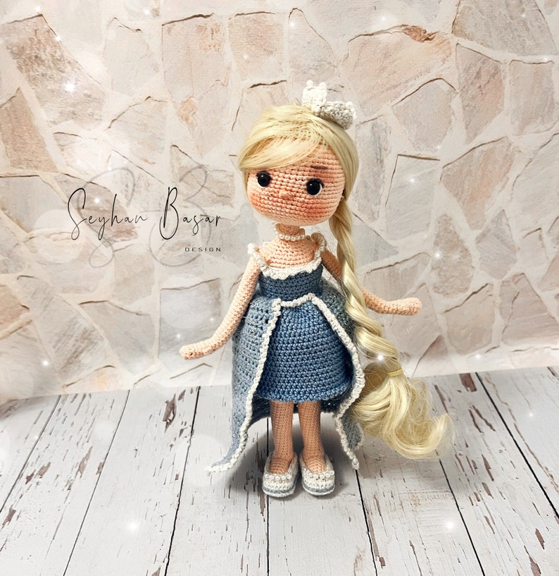 May include: A crocheted doll with blonde hair wearing a blue dress and a crown. The doll is standing on a wooden surface.