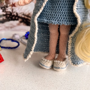 May include: A crocheted doll wearing a blue dress and a white-trimmed robe. The doll has blonde hair and is wearing white shoes with pearl accents.