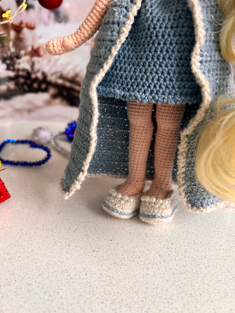 May include: A crocheted doll wearing a blue dress and a white-trimmed robe. The doll has blonde hair and is wearing white shoes with pearl accents.