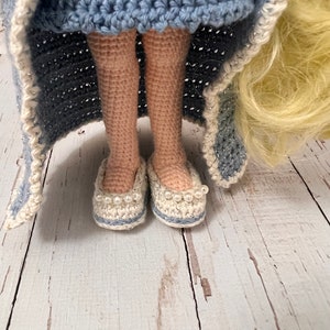 May include: Crocheted doll wearing a blue dress and white crocheted shoes with pearl accents. The doll has long blonde hair and is standing on a white wooden surface.