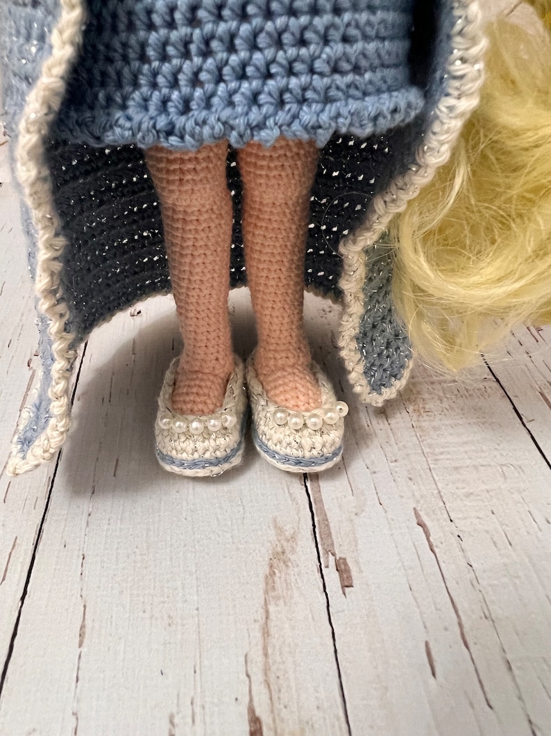 May include: Crocheted doll wearing a blue dress and white crocheted shoes with pearl accents. The doll has long blonde hair and is standing on a white wooden surface.