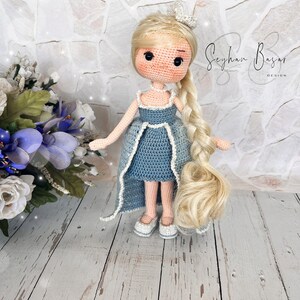 May include: A crocheted doll with long blonde hair wearing a blue dress and white shoes. The doll is standing on a white wooden surface. The text "Seyhan Basar Design" is visible in the background.