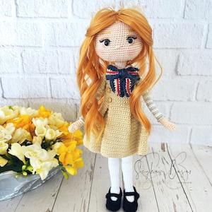 May include: A crocheted doll with long, curly, orange hair, wearing a beige dress, a blue and red bow, white tights, and black shoes. The doll is standing on a wooden surface with a bouquet of yellow flowers in a metal vase.