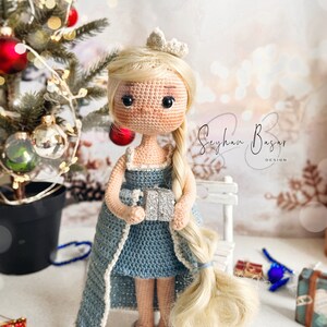 May include: A crocheted doll wearing a blue dress and a crown, holding a silver gift box. The doll has long blonde hair and is standing in front of a white bench and a Christmas tree.
