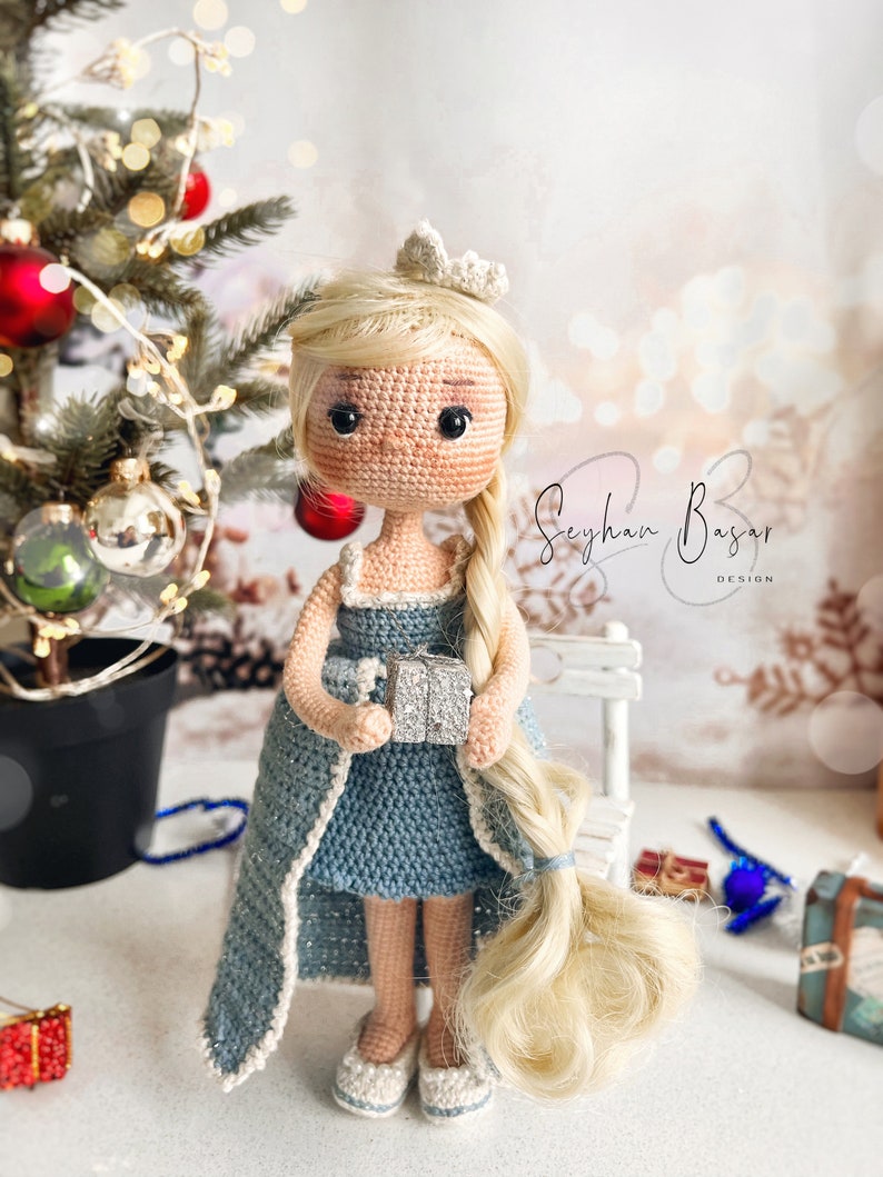 May include: A crocheted doll wearing a blue dress and a crown, holding a silver gift box. The doll has long blonde hair and is standing in front of a white bench and a Christmas tree.