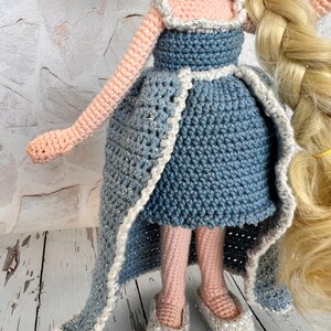 May include: A crocheted doll wearing a blue dress with a white trim and white pearl necklace. The doll has long blonde hair in a braid and is wearing white shoes with pearl accents.