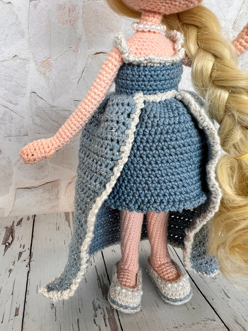 May include: A crocheted doll wearing a blue dress with a white trim and white pearl necklace. The doll has long blonde hair in a braid and is wearing white shoes with pearl accents.