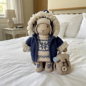 May include: A knitted bunny doll wearing a navy blue hooded jacket with faux fur trim, a matching hat, and a sweater with a snowflake pattern. The doll has a small backpack with a compass charm. The bunny is wearing matching boots.