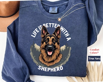 May include: A navy blue crewneck sweatshirt featuring a graphic of a German Shepherd dog head centered above the text "LIFE IS BETTER WITH A SHEPHERD" in white. The design includes a mountain and forest silhouette background.