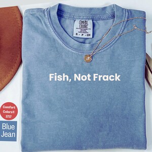 May include: Blue jean Comfort Colors t-shirt with the text "Fish, Not Frack" in white. A gold necklace with a pendant rests on the shirt. A brown hat and dried plant stems are visible in the background.