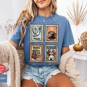 May include: Blue jean Comfort Colors t-shirt featuring four postage stamp designs. The stamps depict a dove, coffee cup, stack of books, and a Cavalier King Charles Spaniel. The shirt is a comfortable, casual style.