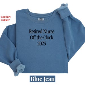May include: Blue jean-colored sweatshirt with the text "Retired Nurse Off the Clock 2025." The sweatshirt is a crew neck style with long sleeves and a soft, textured appearance. The words "Comfort Colors" are in the upper left corner.