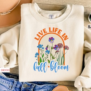 May include: A cream-colored sweatshirt with the text "Live Life in Full Bloom" in orange and blue lettering. The text is surrounded by a colorful floral design.