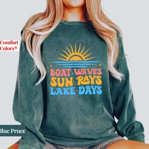 May include: Blue Pruce sweatshirt with a graphic of a sun, waves, and the words "Boat Waves Sun Rays Lake Days" in orange, pink, and blue. The sweatshirt is a Comfort Colors brand.