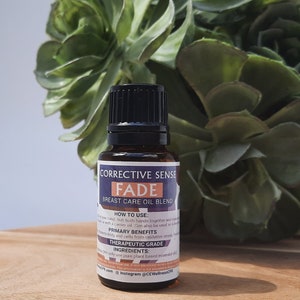 以下が含まれることがあります： 黒いキャップが付いた小さな茶色のガラス瓶で、「Corrective Sense Fade Breast Care Oil Blend」とラベル付けされています。瓶は木の表面に置かれ、背景には緑の植物があります。ラベルには、使用方法、主な利点、成分が記載されています。
