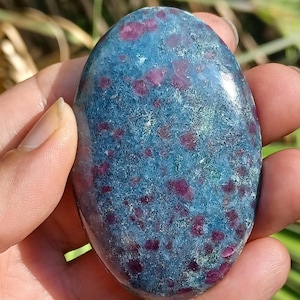May include: Oval-shaped gemstone with a blue-green base color and scattered red inclusions. The polished stone is held in a hand, with a blurred green background.