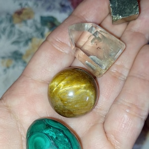 May include: Assortment of polished gemstones including a clear quartz crystal, a golden tiger's eye sphere, a green malachite oval, and a pyrite cube. These stones are often used for their metaphysical properties.