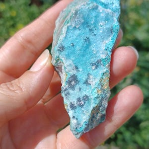 May include: A vibrant turquoise mineral specimen with dark gray and black inclusions. The rough-hewn stone displays a crystalline structure and varying shades of blue and green. The natural rock is held in a hand, showcasing its texture and color.