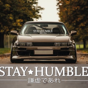 May include: A brown car with a windshield banner that reads "STAY HUMBLE" in white text with a star between the words. The banner also has Japanese text below the words. The car is parked on a paved road with trees on either side.