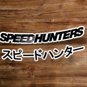 Może przedstawiać: Biała naklejka z czarnym napisem "SPEEDHUNTERS" nad japońskimi znakami. Naklejka umieszczona na drewnianej powierzchni.