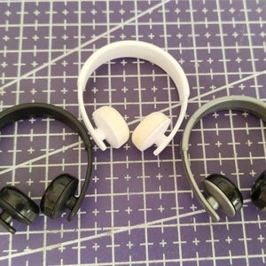 Könnte beinhalten: Drei Paare On-Ear-Kopfhörer in Schwarz, Weiß und Grau. Jedes Paar hat einen gebogenen Kopfbügel und zwei Ohrmuscheln. Die Kopfhörer sind auf einer violetten Rasteroberfläche angeordnet.