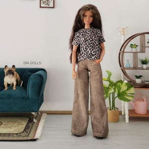 Op de afbeelding: Een modepop met lang bruin haar draagt een top met korte mouwen met luipaardprint en een wijde bruine spijkerbroek. De pop heeft beweegbare gewrichten en staat rechtop. De tekst DIA DOLLS is zichtbaar op de muur achter de pop.
