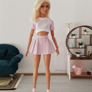 May include: A fashion doll with long blonde hair wearing a white and light pink raglan crop top, a pleated light pink mini skirt with two silver buttons, light pink ankle socks, and white trainers. The text DIA DOLLS is visible on the wall.
