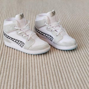 Può includere: Un paio di sneakers alte bianche con un motivo a stelle e quadretti sul lato. Le sneakers hanno lacci bianchi e una suola azzurra. Sulla linguetta della scarpa ci sono le lettere "MF".