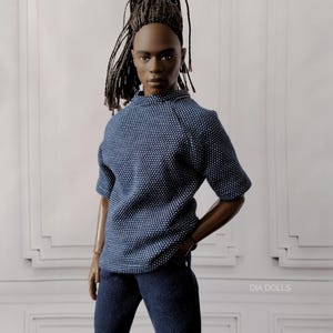 Può includere: Bambola alla moda con la pelle scura, che indossa una camicia blu a maniche corte fantasia e jeans blu scuro. La bambola ha i capelli intrecciati scuri ed è posata su uno sfondo bianco. Il testo "DIA DOLLS" è visibile.