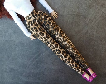 May include: A doll wearing a white long-sleeved shirt and brown and black leopard print pants. The doll is wearing pink high heels.