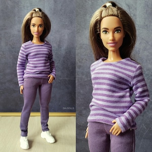 Peut inclure: Une poupée vêtue d'un haut à manches longues rayé violet et blanc et d'un pantalon violet assorti. La poupée porte également des baskets blanches. La poupée a de longs cheveux bruns et se tient devant un fond gris.