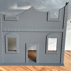 以下が含まれることがあります： レンガ模様のグレーのミニチュアの建物のファサード。 ファサードには、2つの長方形の窓、中央のドア、三角形の屋根部分があります。 建物は頑丈な素材でできており、模型製作や展示用に設計されています。