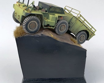 May include: A green military model truck with a flatbed trailer, on a black and brown display base. The truck is driving up a small hill of brown sand.