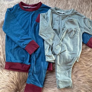 May include: Two sets of long-sleeved shirts and trousers. One set is deep blue with dark red cuffs and hem. The second set is sage green with matching cuffs and hem. Both outfits feature crew necklines and elasticated waistbands on the trousers.