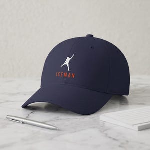 May include: A navy blue baseball cap featuring a white silhouette graphic of a person reaching upward with one arm extended. Below the graphic, the word ICEMAN is printed in orange capital letters. The cap has a curved brim and a structured crown.