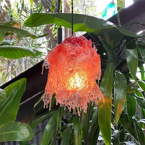 Op de afbeelding: Een rode, oranje en gele glazen hanglamp met een druppelend, gesmolten wasontwerp. De lamp hangt aan een zwart snoer en is omgeven door groene planten.