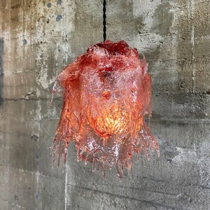 Op de afbeelding: Een roze glazen hanglamp met een druppelvormig, gesmolten ontwerp. De lamp hangt aan een zwart snoer tegen een betonnen muur.