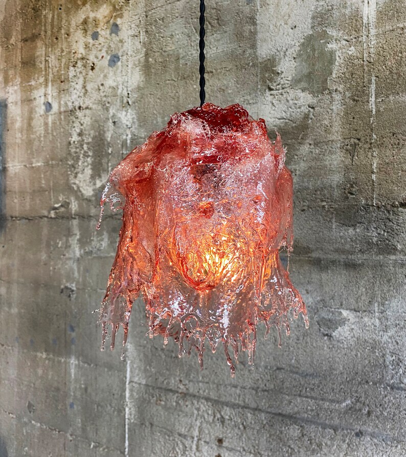 Op de afbeelding: Een roze glazen hanglamp met een druppelvormig, gesmolten ontwerp. De lamp hangt aan een zwart snoer tegen een betonnen muur.