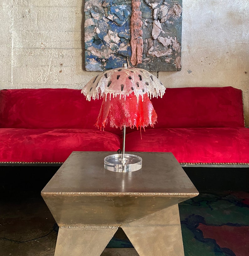 May include: A red and white lamp with a unique dripping design. The lamp is on a silver table with a geometric shape.