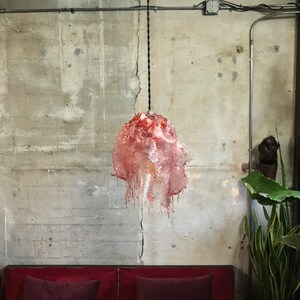 Op de afbeelding: Een rode en roze glazen hanglamp hangt aan een zwart snoer tegen een betonnen muur. De lamp heeft een druppelende, gesmolten uitstraling.