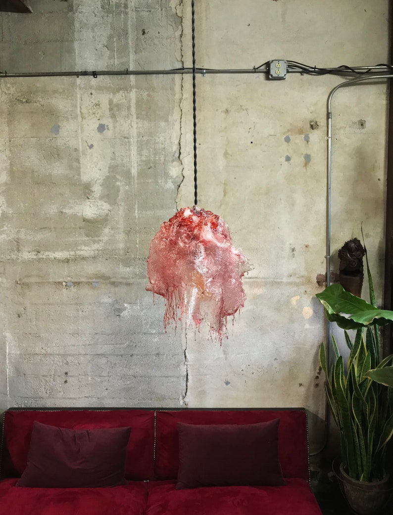 Op de afbeelding: Een rode en roze glazen hanglamp hangt aan een zwart snoer tegen een betonnen muur. De lamp heeft een druppelende, gesmolten uitstraling.