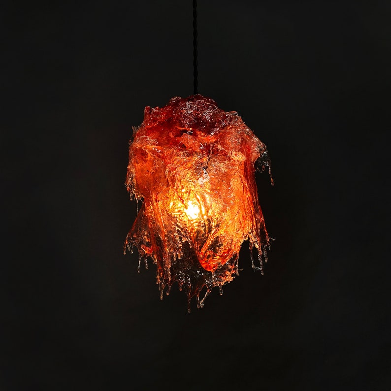 Op de afbeelding: Een hanglamp gemaakt van oranje hars die lijkt op druipende lava. De lamp is verlicht en geeft een warm licht.