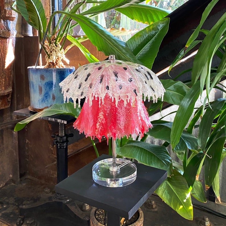 May include: A pink and white lamp with a unique, dripping design. The lamp is on a black stand and has a clear base.