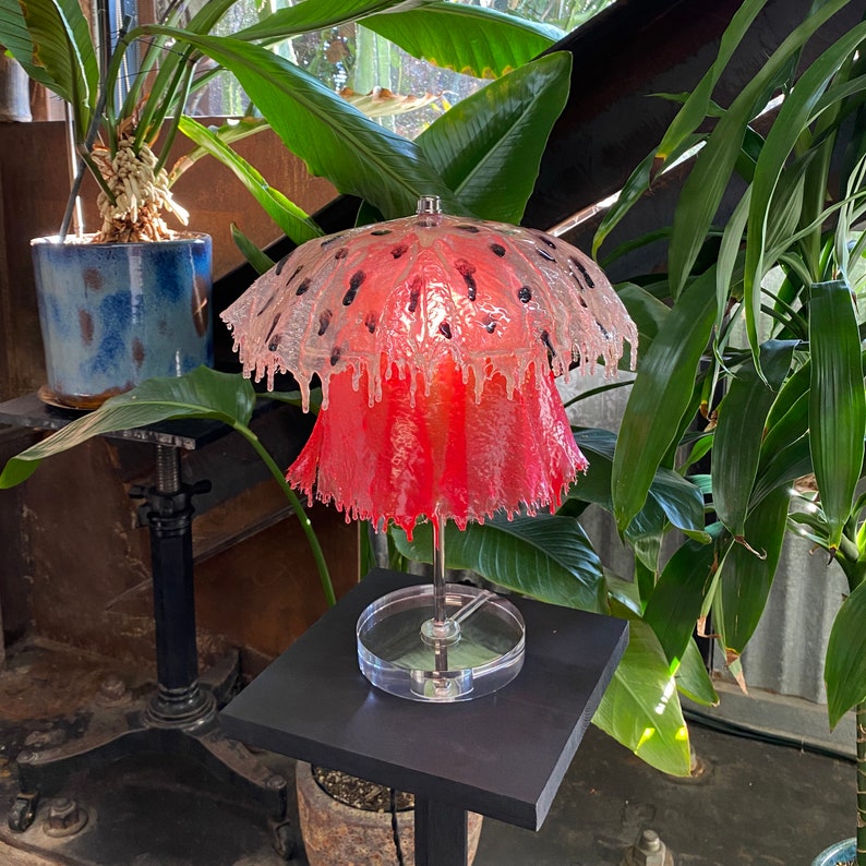 May include: A pink and white glass lamp with a unique, organic shape resembling a flower. The lamp is on a black pedestal base and has a clear glass shade.