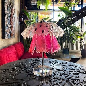 May include: A pink and clear glass lampshade with a unique dripping design. The lampshade is on a black metal base with a clear glass bottom. The lamp is sitting on a dark wooden table with intricate carvings.