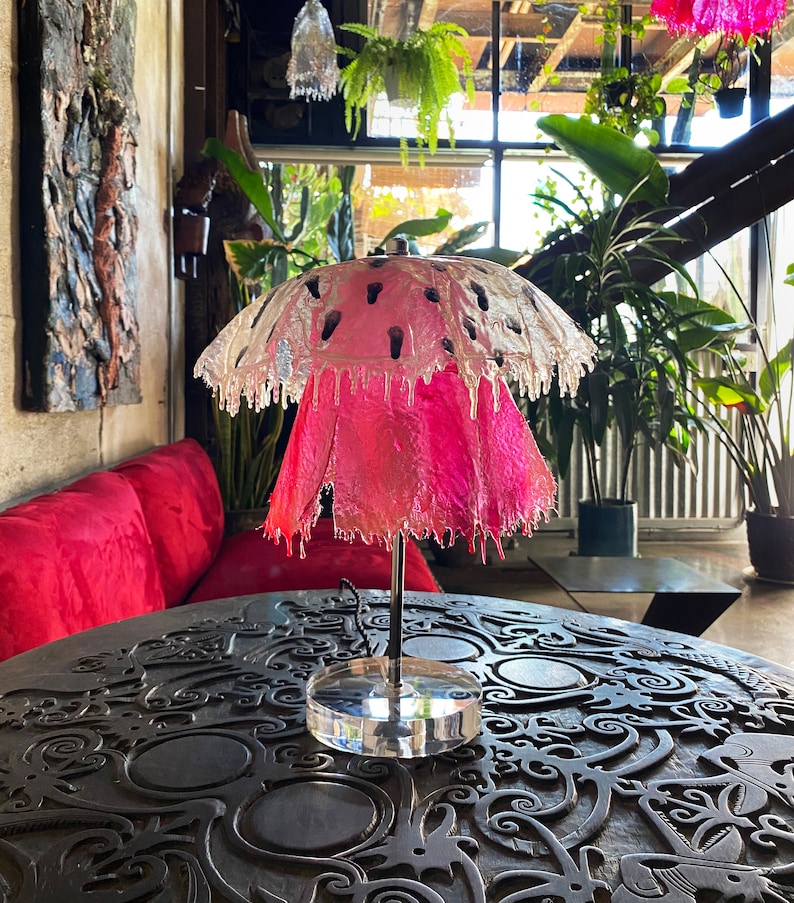 May include: A pink and clear glass lampshade with a unique dripping design. The lampshade is on a black metal base with a clear glass bottom. The lamp is sitting on a dark wooden table with intricate carvings.