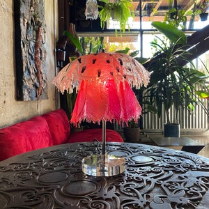 May include: A pink and clear glass lamp shaped like a mushroom with a clear acrylic base. The lamp is sitting on a dark wooden table with intricate carvings.