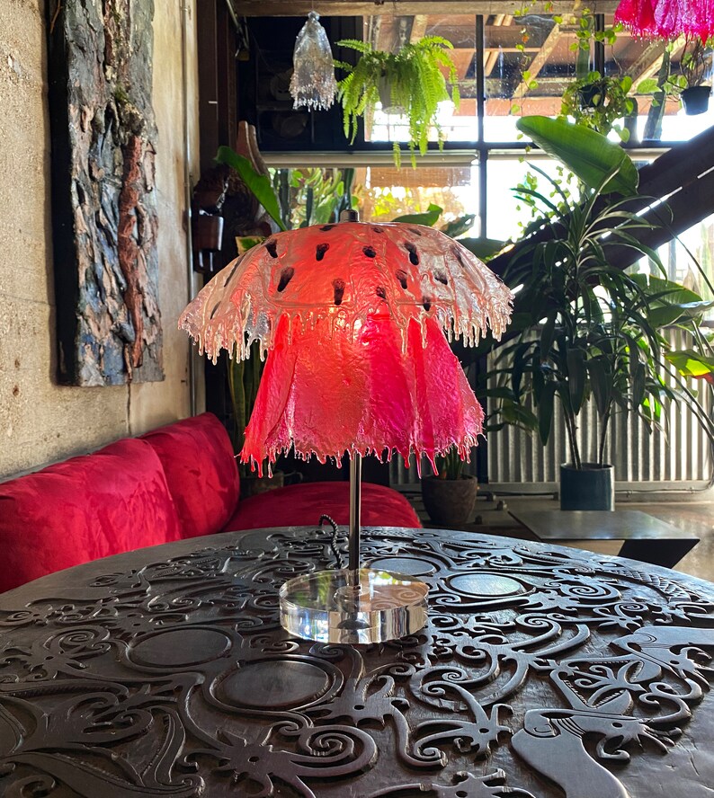 May include: A pink and clear glass lamp shaped like a mushroom with a clear acrylic base. The lamp is sitting on a dark wooden table with intricate carvings.