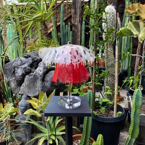 May include: A red and white lamp with a mushroom-shaped shade. The lamp is on a black stand and is surrounded by plants.