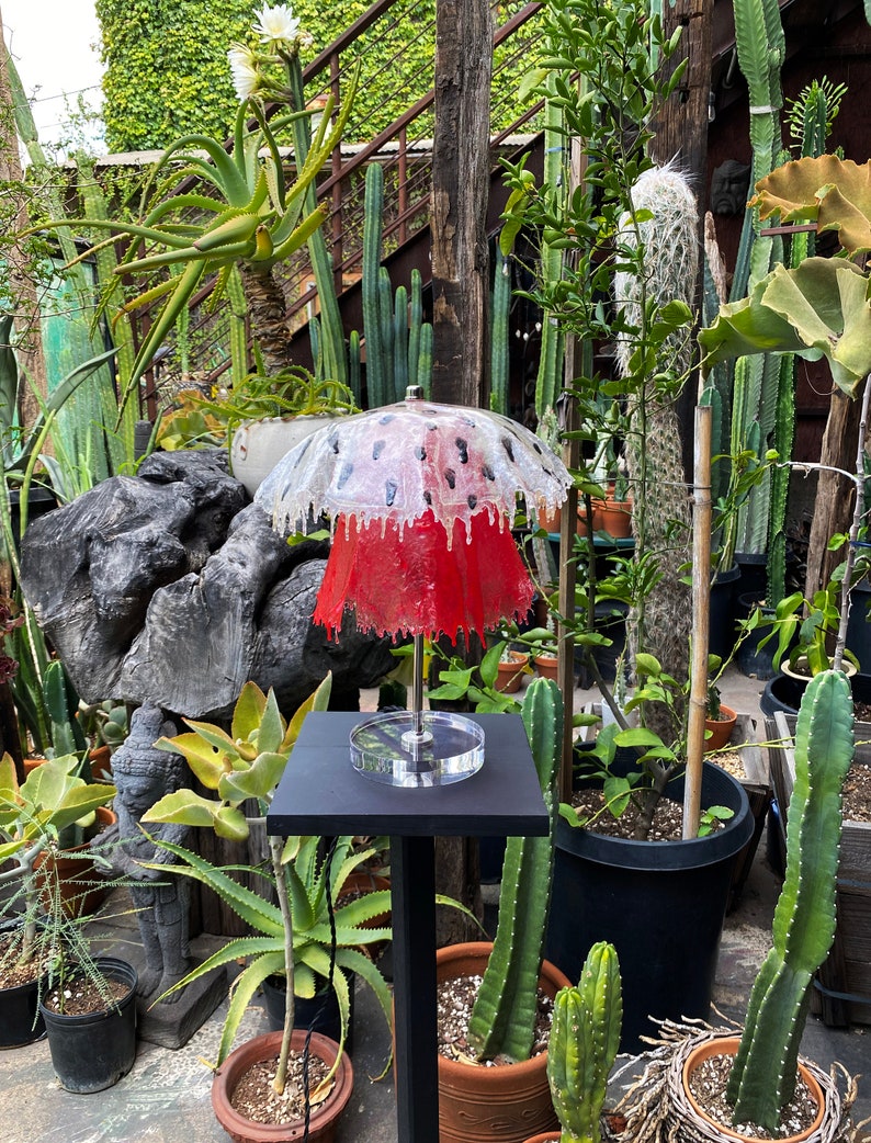 May include: A red and white lamp with a mushroom-shaped shade. The lamp is on a black stand and is surrounded by plants.